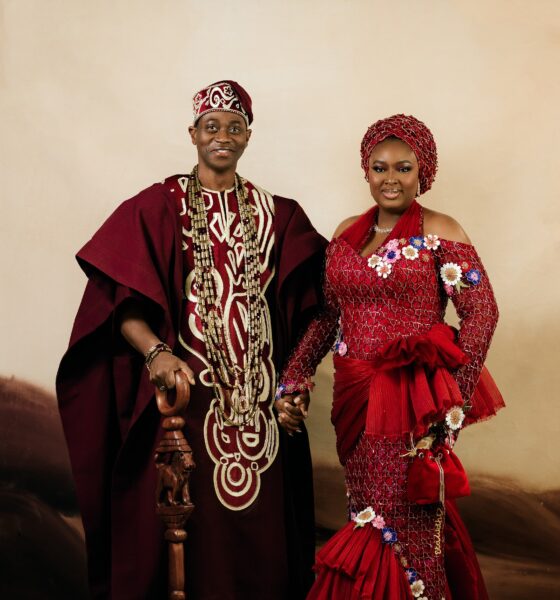 Lateef Adedimeji holding a carved wooden lion staff while holding hands with his wife Mo Bimpe in matching red traditional Yoruba attire.