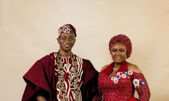 Lateef Adedimeji holding a carved wooden lion staff while holding hands with his wife Mo Bimpe in matching red traditional Yoruba attire.