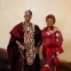 Lateef Adedimeji holding a carved wooden lion staff while holding hands with his wife Mo Bimpe in matching red traditional Yoruba attire.