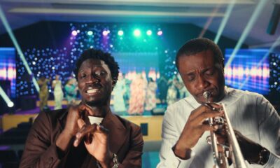 Greatman Takit in a brown suit singing beside Nathaniel Bassey playing his signature silver trumpet in white traditional attire on a brightly lit stage.