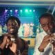 Greatman Takit in a brown suit singing beside Nathaniel Bassey playing his signature silver trumpet in white traditional attire on a brightly lit stage.