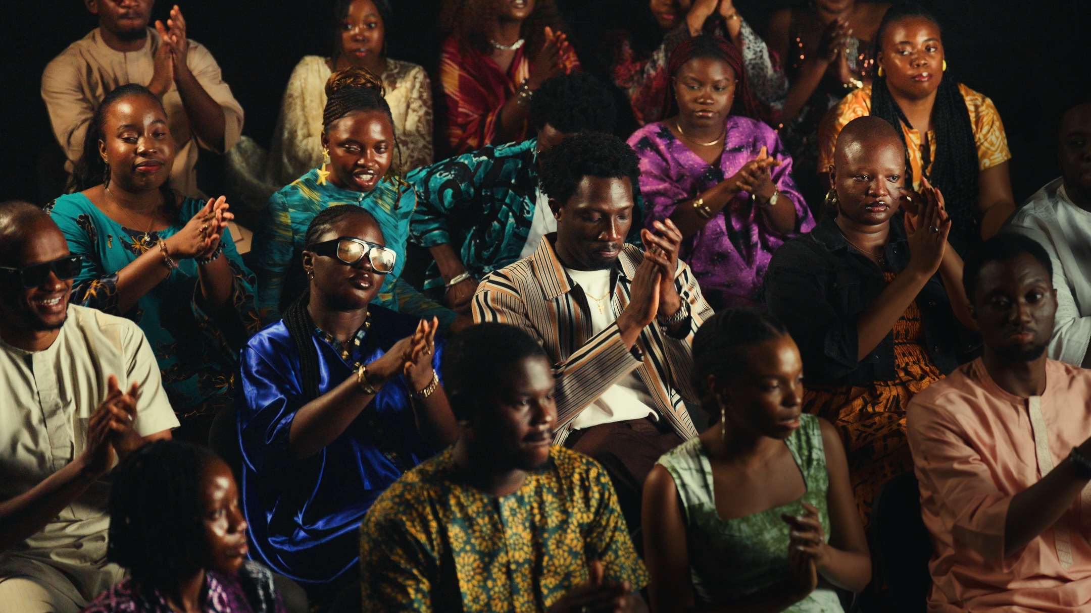 Greatman Takit sitting among a choir and audience clad in colorful African traditional wear as they clap and worship together in an auditorium.