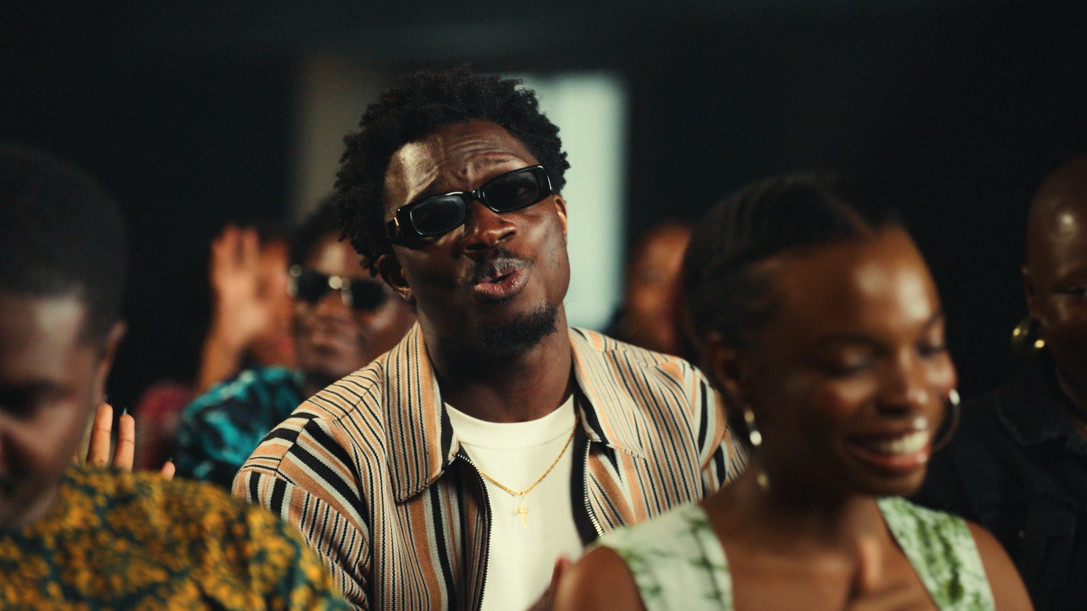 Gospel music minister Greatman Takit wearing a striped jacket and dark sunglasses, singing in an auditorium during the "You Are Good" music video shoot.
