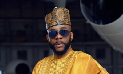 : Ebuka Obi-Uchendu wearing a metallic gold Babariga with geometric embroidery, a patterned Hula cap, and aviator sunglasses inside an airplane hangar.