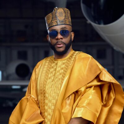: Ebuka Obi-Uchendu wearing a metallic gold Babariga with geometric embroidery, a patterned Hula cap, and aviator sunglasses inside an airplane hangar.