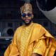 : Ebuka Obi-Uchendu wearing a metallic gold Babariga with geometric embroidery, a patterned Hula cap, and aviator sunglasses inside an airplane hangar.
