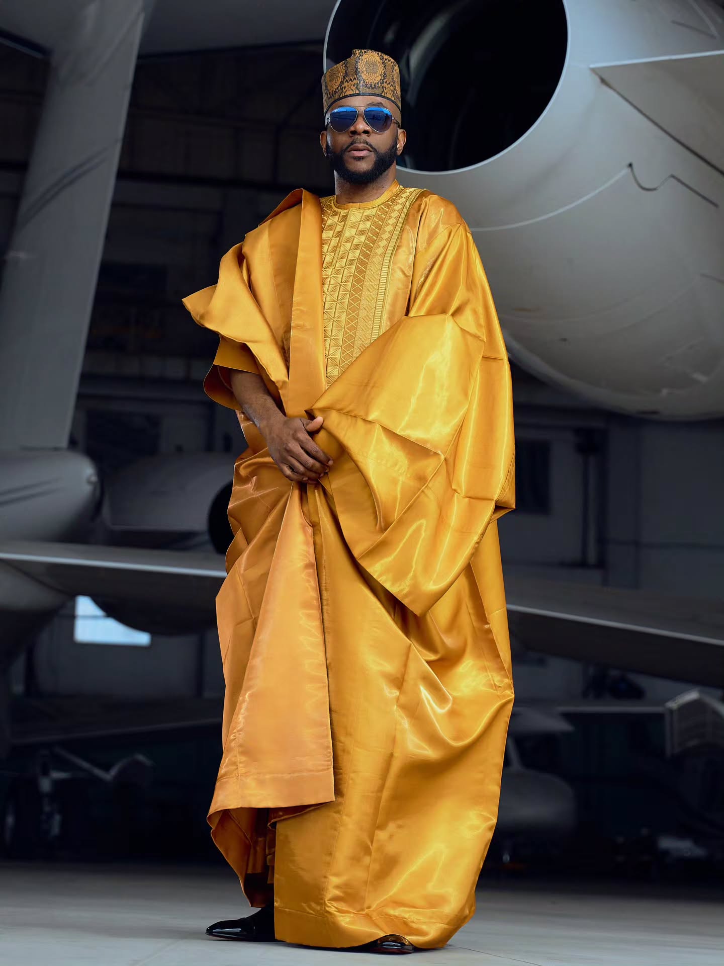 Full-length photo of Ebuka Obi-Uchendu standing in a three-piece gold Babariga robe, python-print cap, and black patent dress shoes next to a private jet.