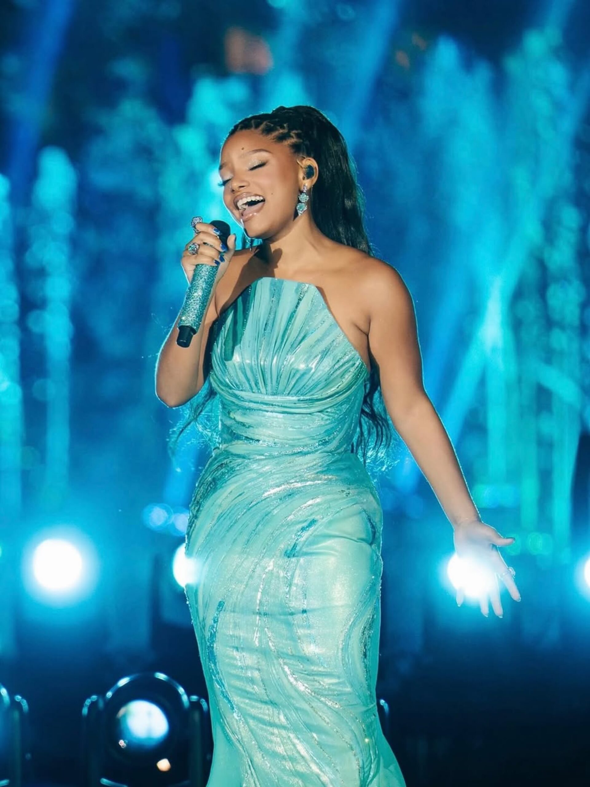 Halle Bailey singing on stage in a metallic teal gown holding a teal microphone.