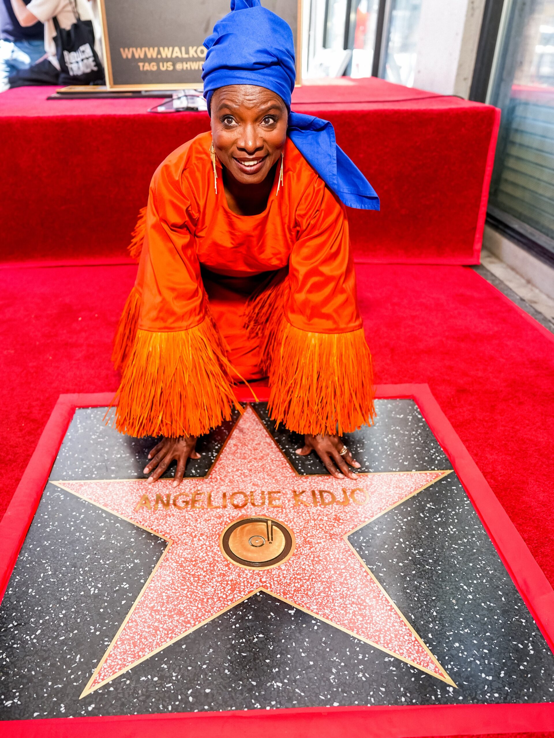 Angélique Kidjo kneeling with hands on her Hollywood Walk of Fame star in an orange outfit and blue headwrap