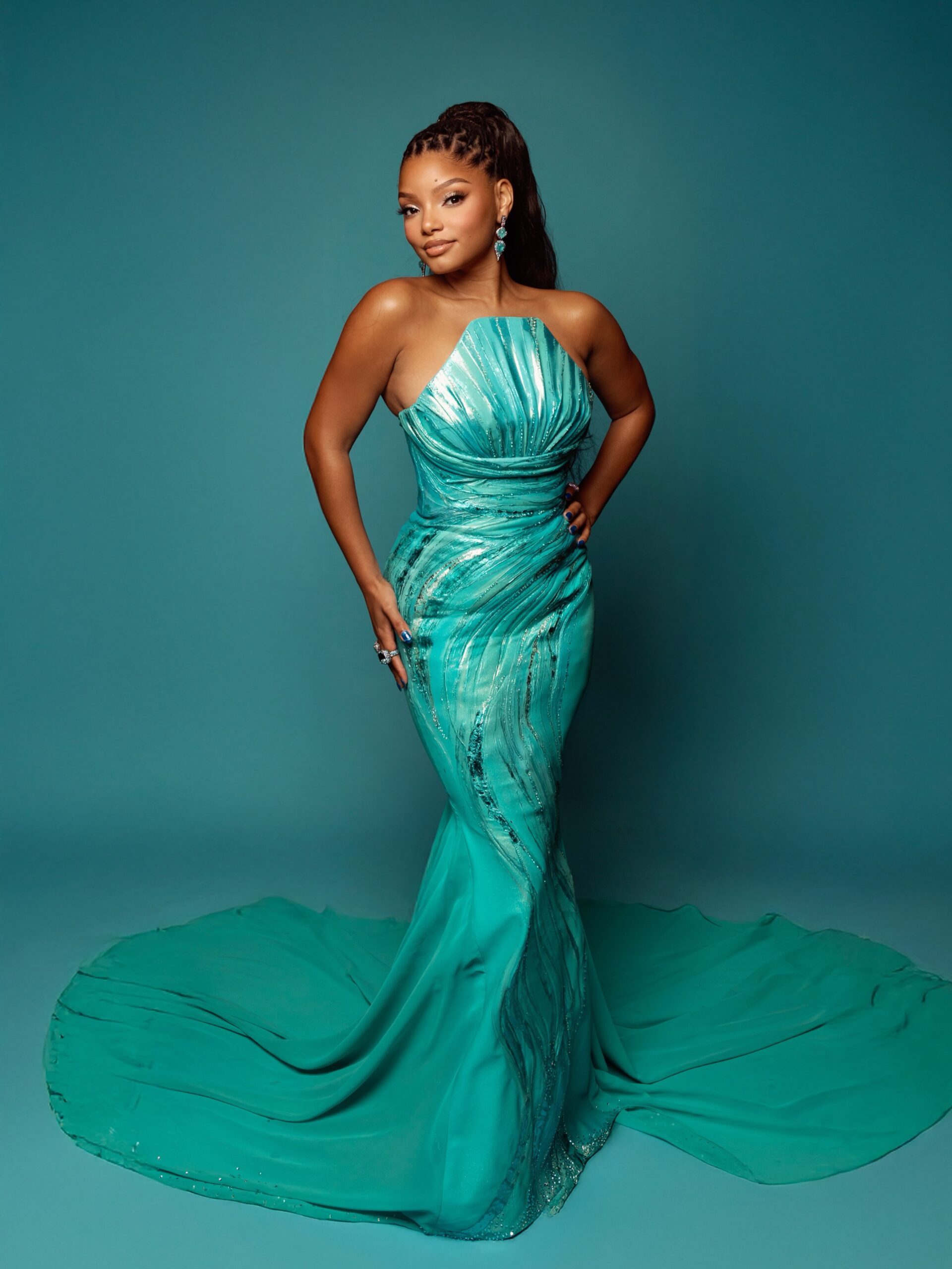 Halle Bailey posing in a full-length metallic teal mermaid gown with sweeping train.
