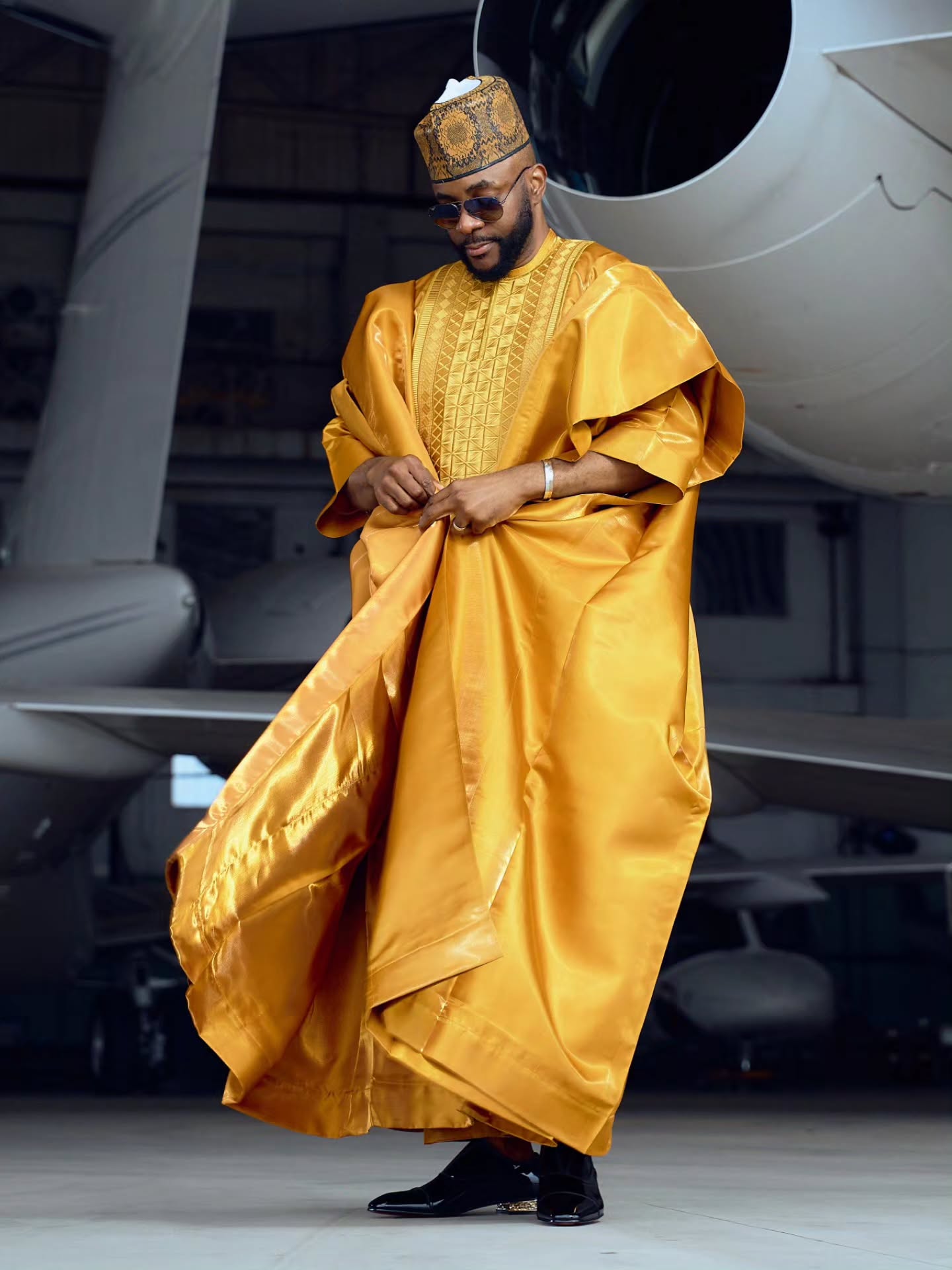 Ebuka Obi-Uchendu adjusting the draped sleeve of his gold Babariga outfit in an aviation hangar.