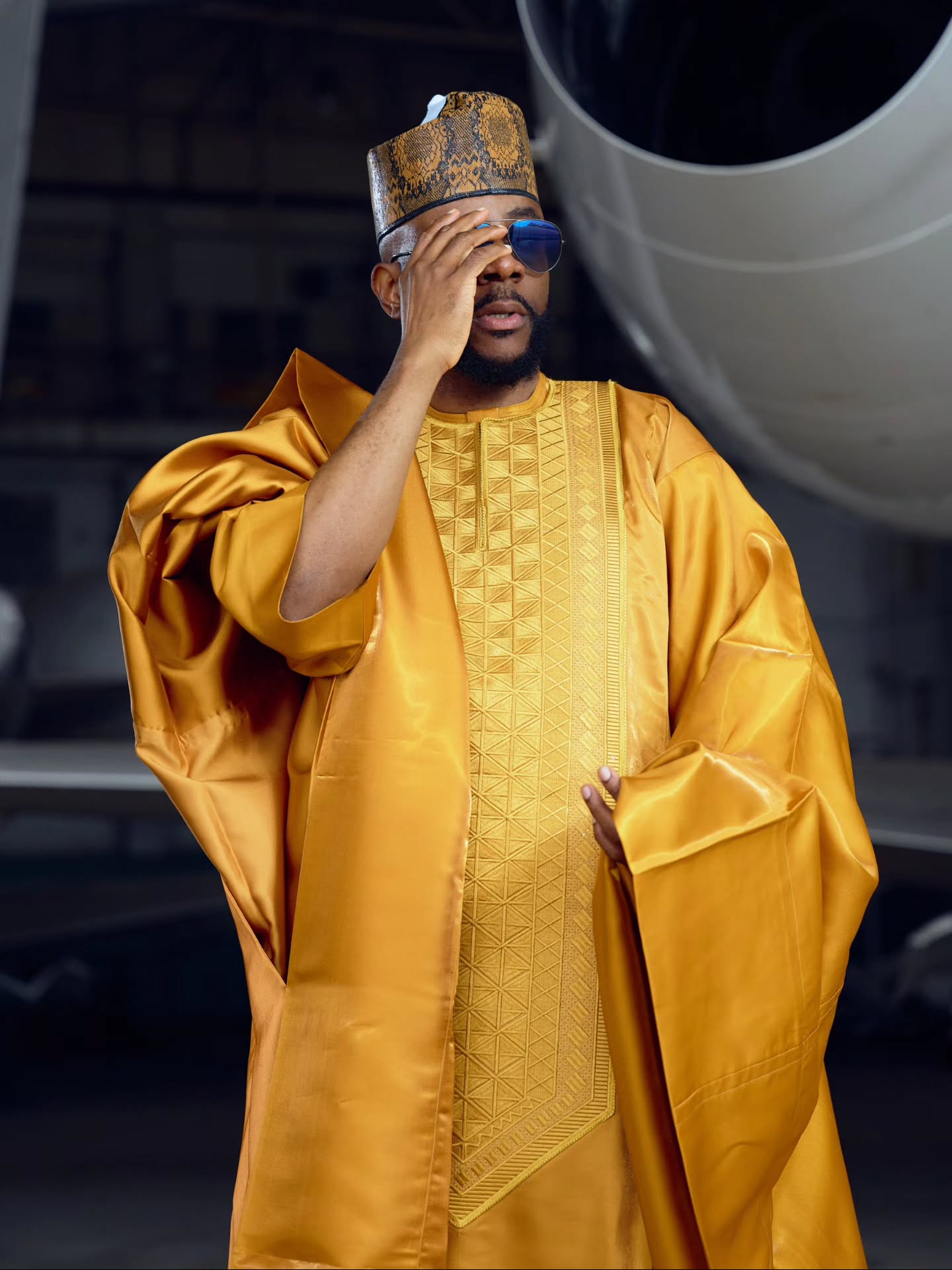 Ebuka Obi-Uchendu posing with one hand touching his sunglasses, wearing a gold embroidered Babariga and patterned cap.