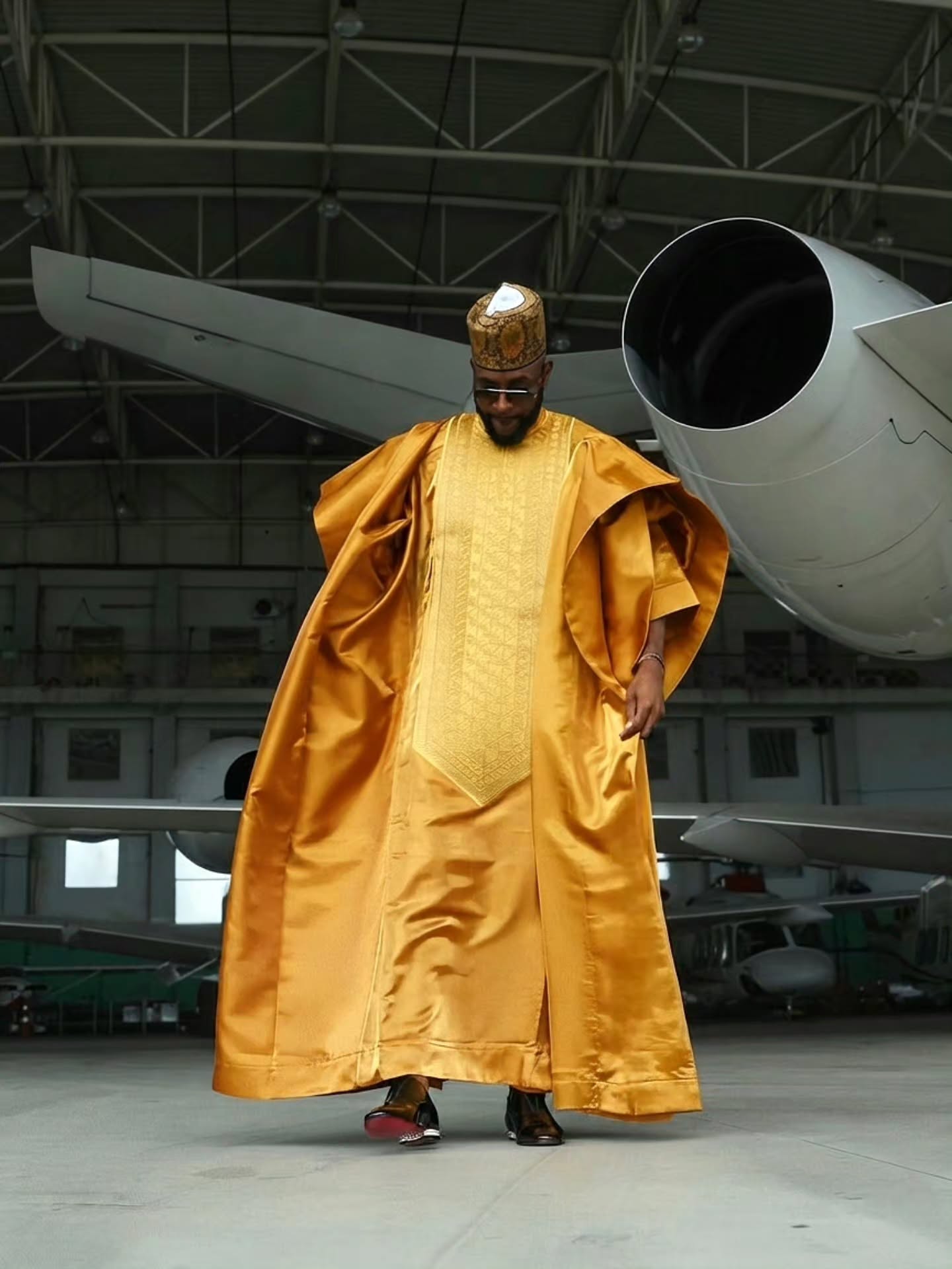 Wide shot of Ebuka Obi-Uchendu walking forward in a flowing gold Babariga inside a private jet hangar.