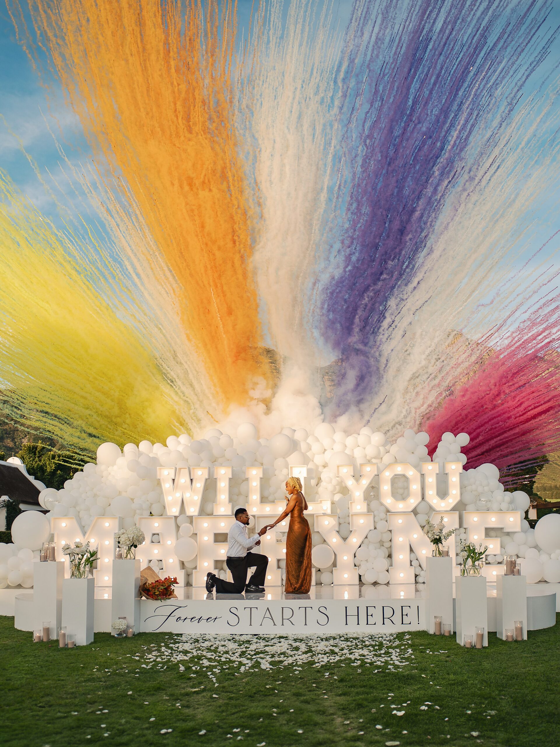 Engagement proposal with colorful smoke cannons, white balloons, and illuminated letters in Cape Town.