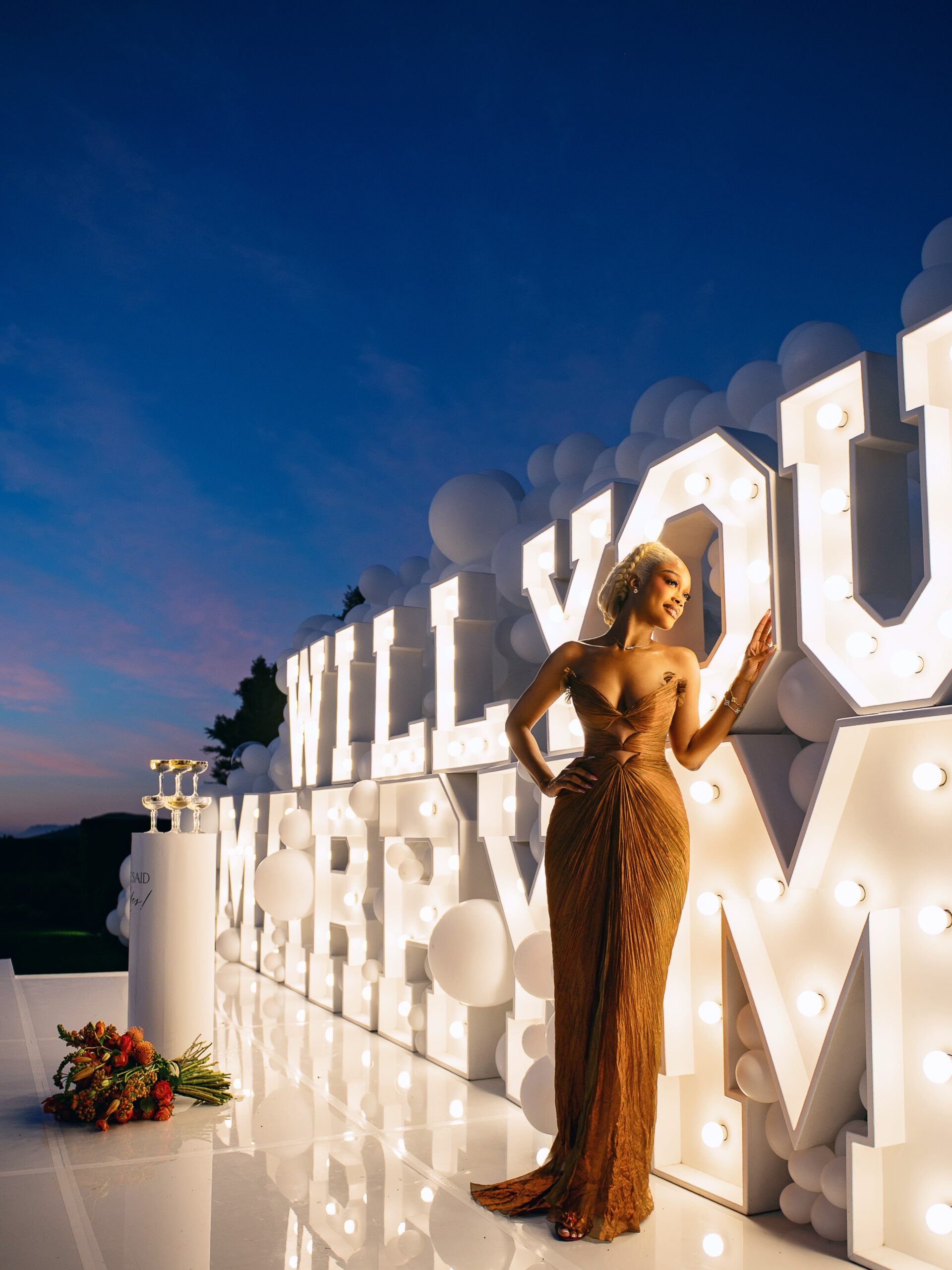 Diana Eneje standing beside illuminated "WILL YOU MARRY ME" marquee letters at dusk.