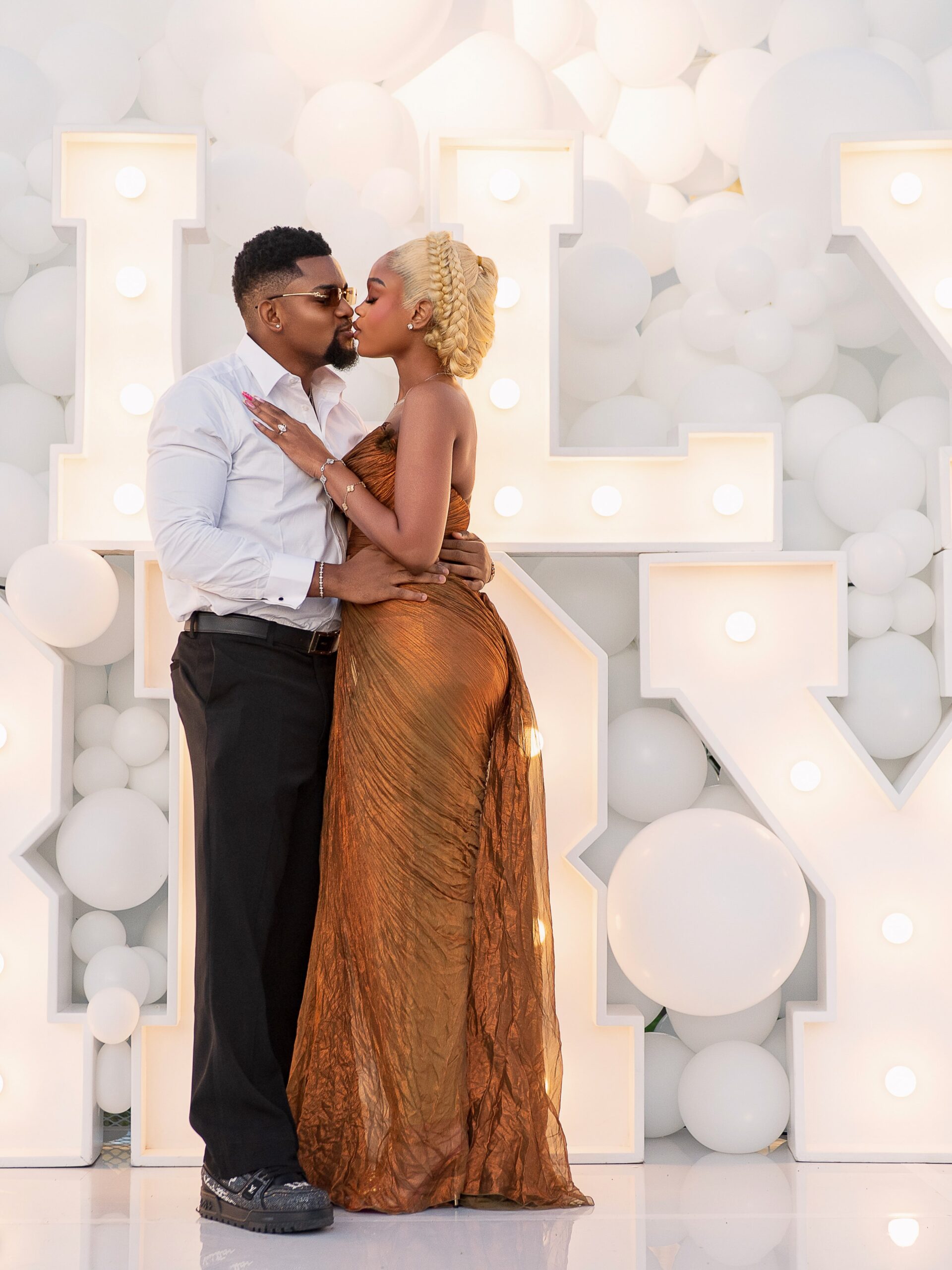 Diana Eneje kissing her fiancé in front of white balloons and illuminated marquee letters.