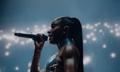 Ayra Starr performing live on stage at All Points East 2026 festival holding a microphone in a metallic silver two-piece outfit with a sleek ponytail.