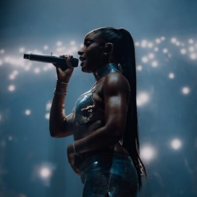Ayra Starr performing live on stage at All Points East 2026 festival holding a microphone in a metallic silver two-piece outfit with a sleek ponytail.