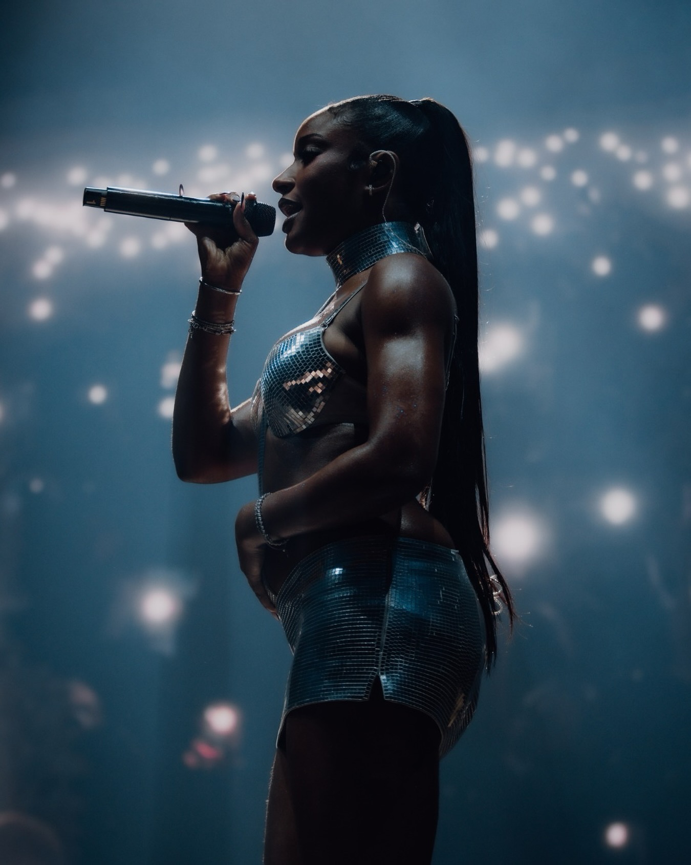 Ayra Starr performing live on stage at All Points East 2026 festival holding a microphone in a metallic silver two-piece outfit with a sleek ponytail.