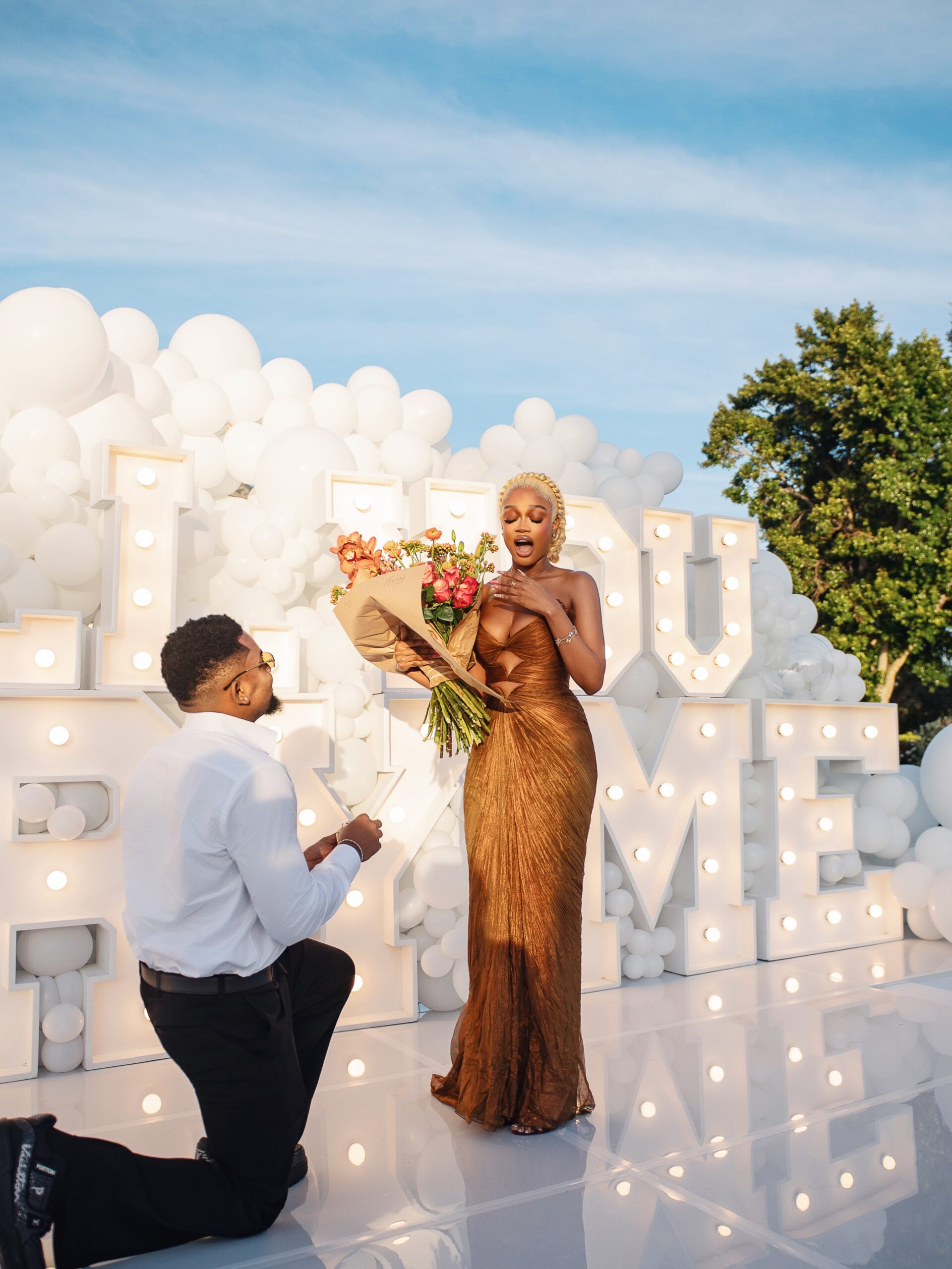 Pascal Godwin on one knee proposing to Diana Eneje holding a large flower bouquet.