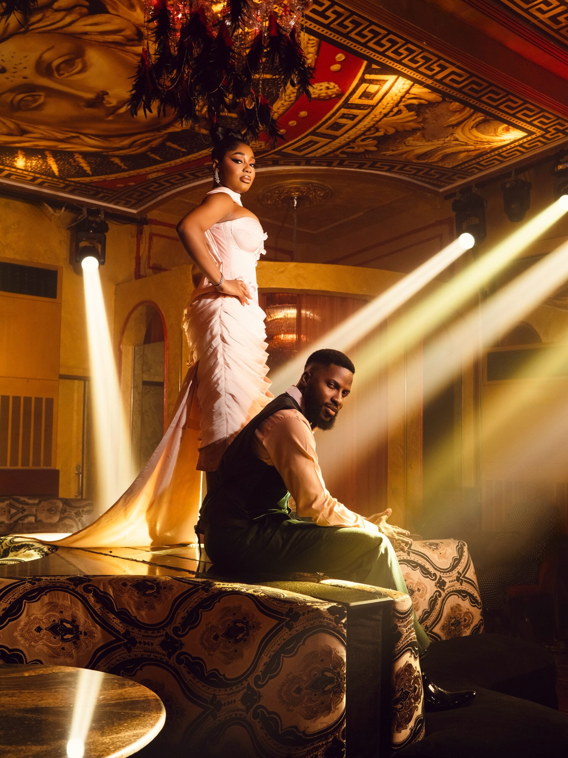 : Kiky Festus standing in a draped pink train gown next to Akin Faminu seated in a green tailored suit under stage lights.