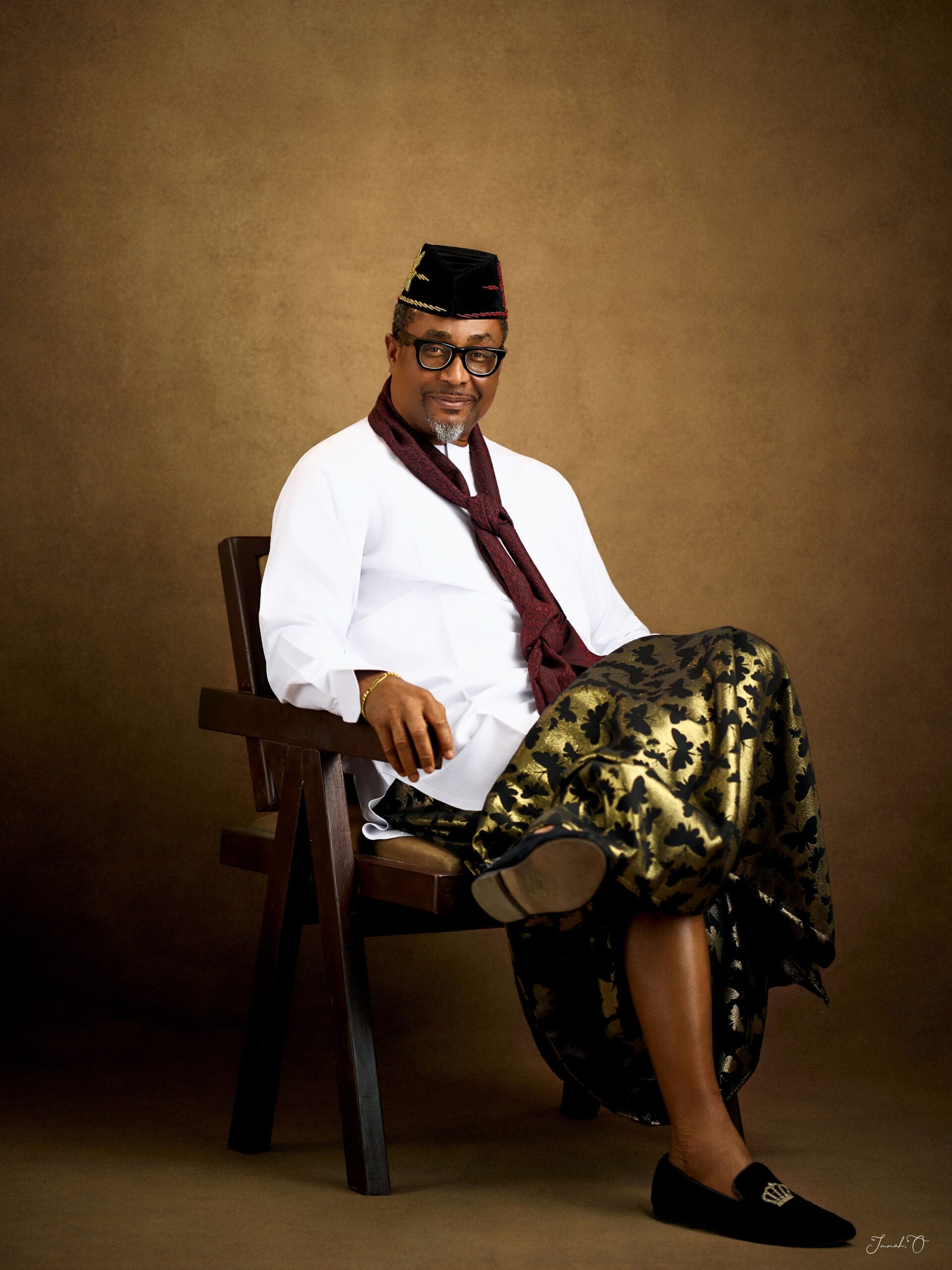 Tony Umez wearing black crown-embroidered loafers, a patterned gold wrapper, and a white tunic.