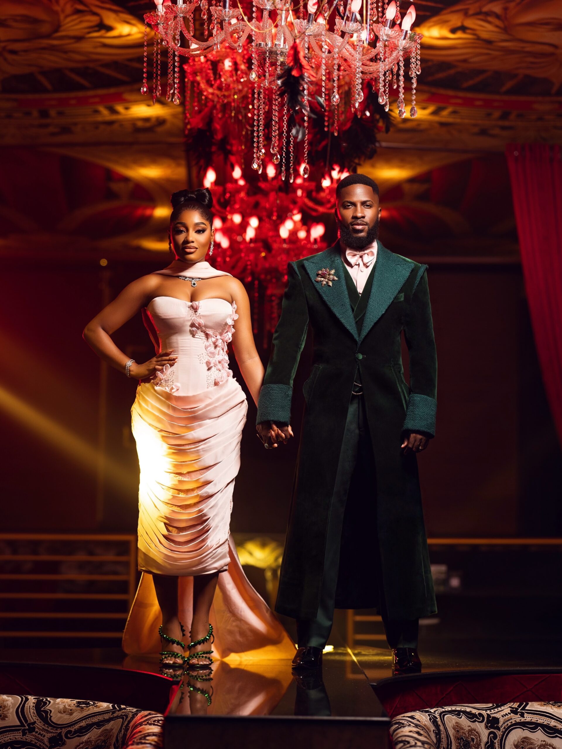 Akin Faminu and Kiky Festus standing together in forest green and blush pink couture under a red chandelier.