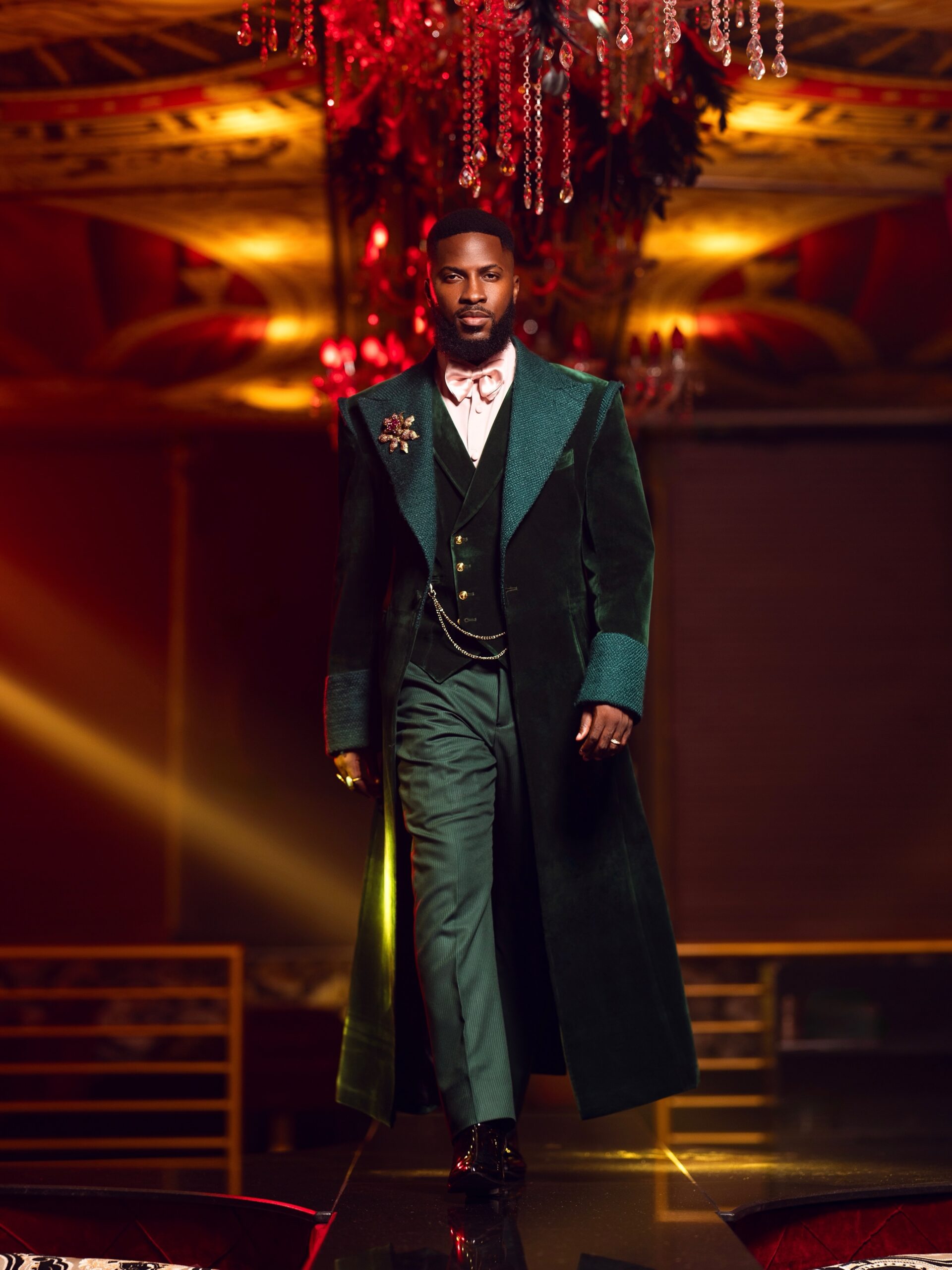 Akin Faminu walking in a dark forest green Deji and Kola velvet trench coat suit with a blush pink bow tie.