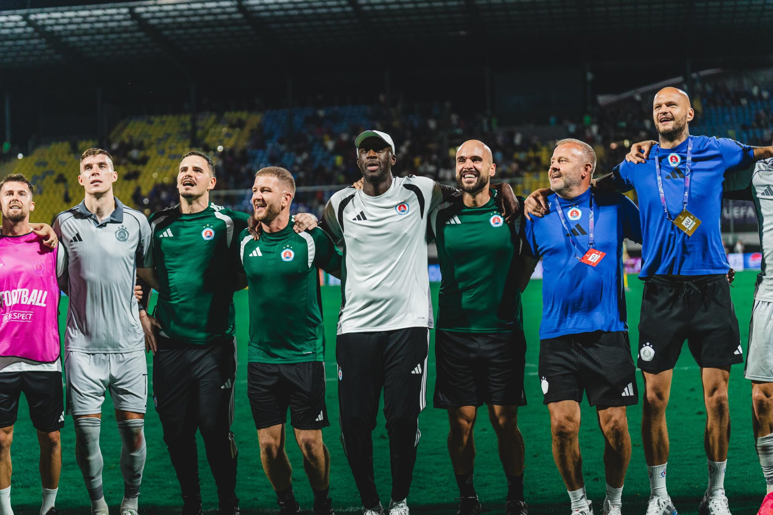Slovan Bratislava players, coaching staff, and manager Yaya Touré standing arm-in-arm on the stadium pitch facing the stands.