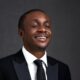 Nathaniel Bassey smiling with eyes closed while wearing a black suit jacket, white shirt, and dark tie for his 45th birthday.