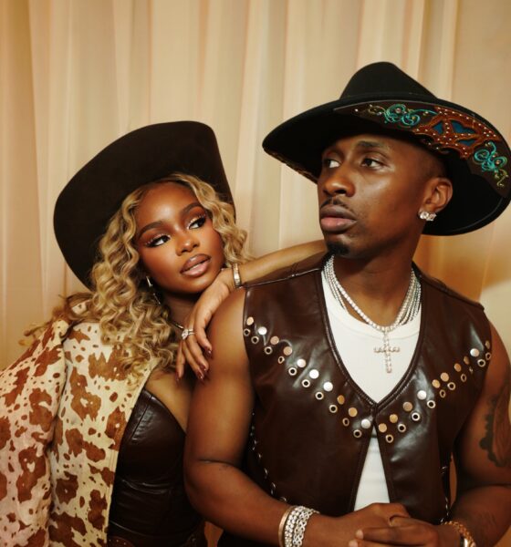 Priscilla Ojo in cow-hide print jacket and leather corset leaning on Juma Jux wearing brown leather vest and decorated cowboy hat.