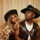 Priscilla Ojo in cow-hide print jacket and leather corset leaning on Juma Jux wearing brown leather vest and decorated cowboy hat.