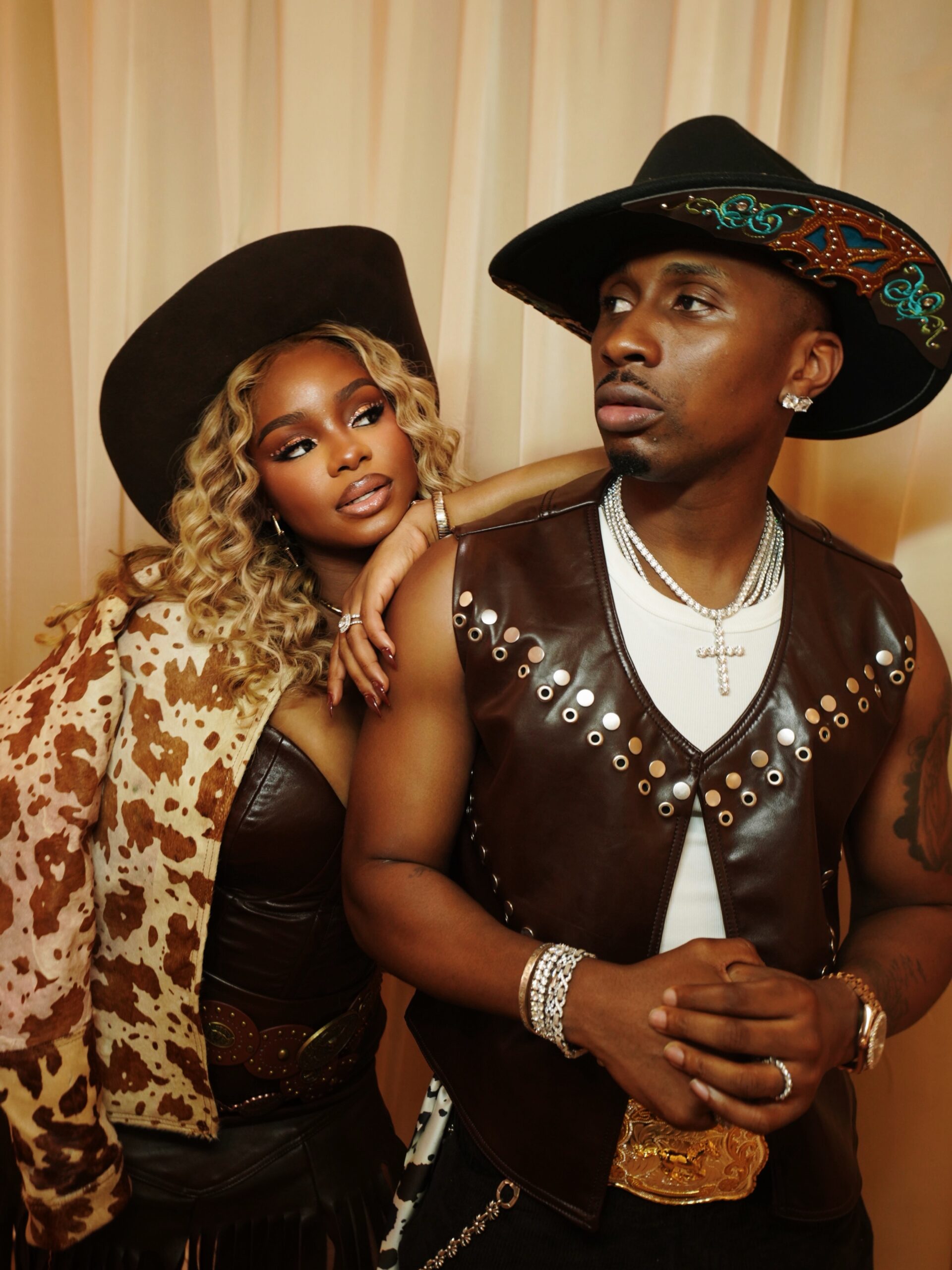 Priscilla Ojo in cow-hide print jacket and leather corset leaning on Juma Jux wearing brown leather vest and decorated cowboy hat.
