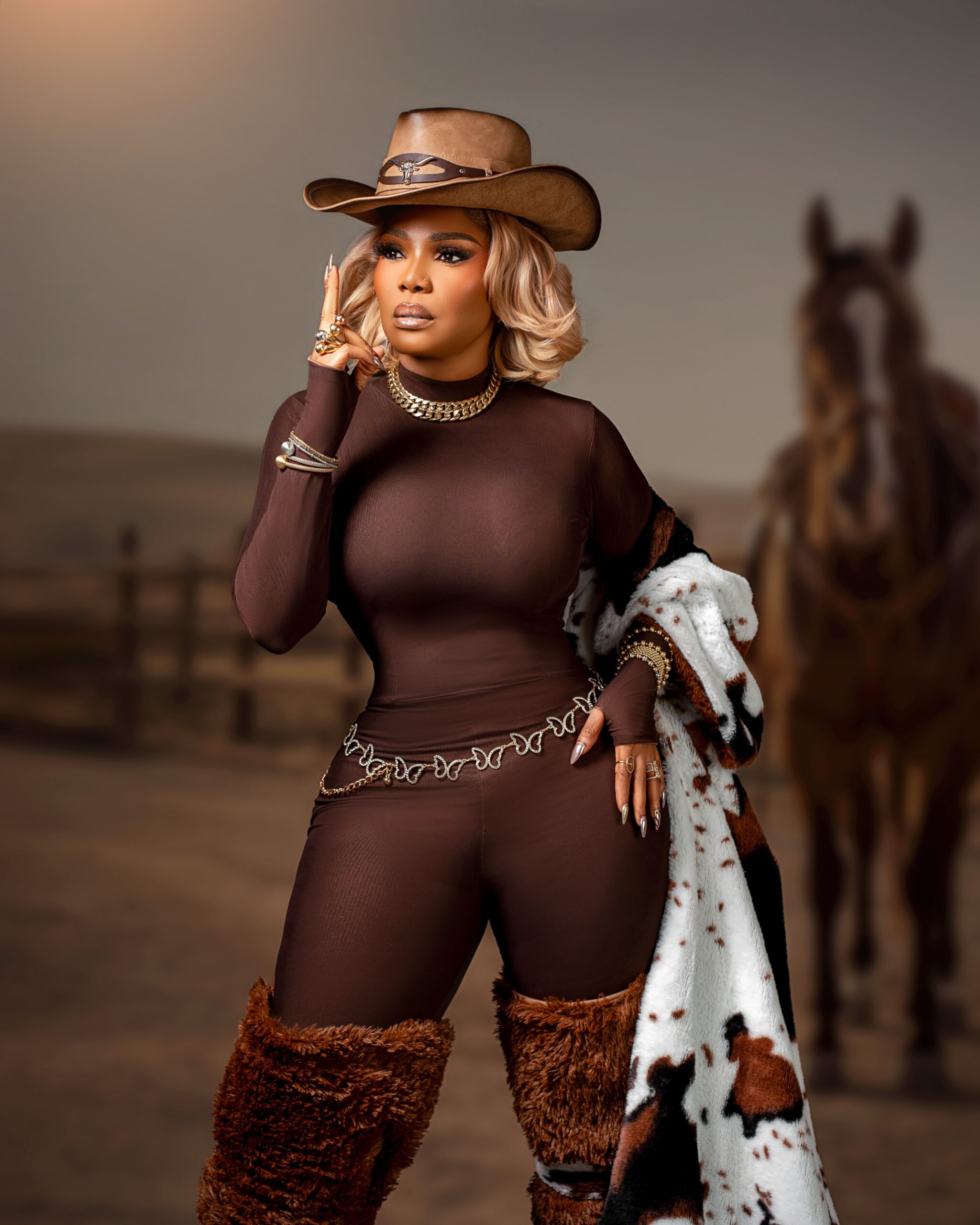 Iyabo Ojo adjusting leather cowboy hat with longhorn emblem, wearing gold Cuban link necklace and honey blonde hair.