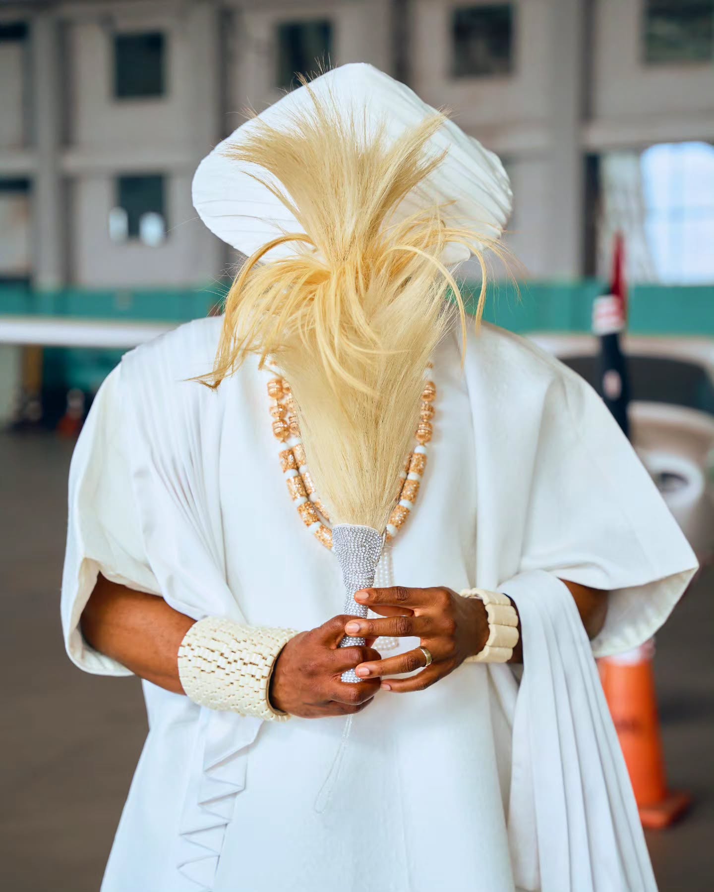Ebuka Obi-Uchendu holding a crystal-embellished horsetail whip while wearing a white Japanese-inspired tunic, coral beads, and woven bead cuffs.