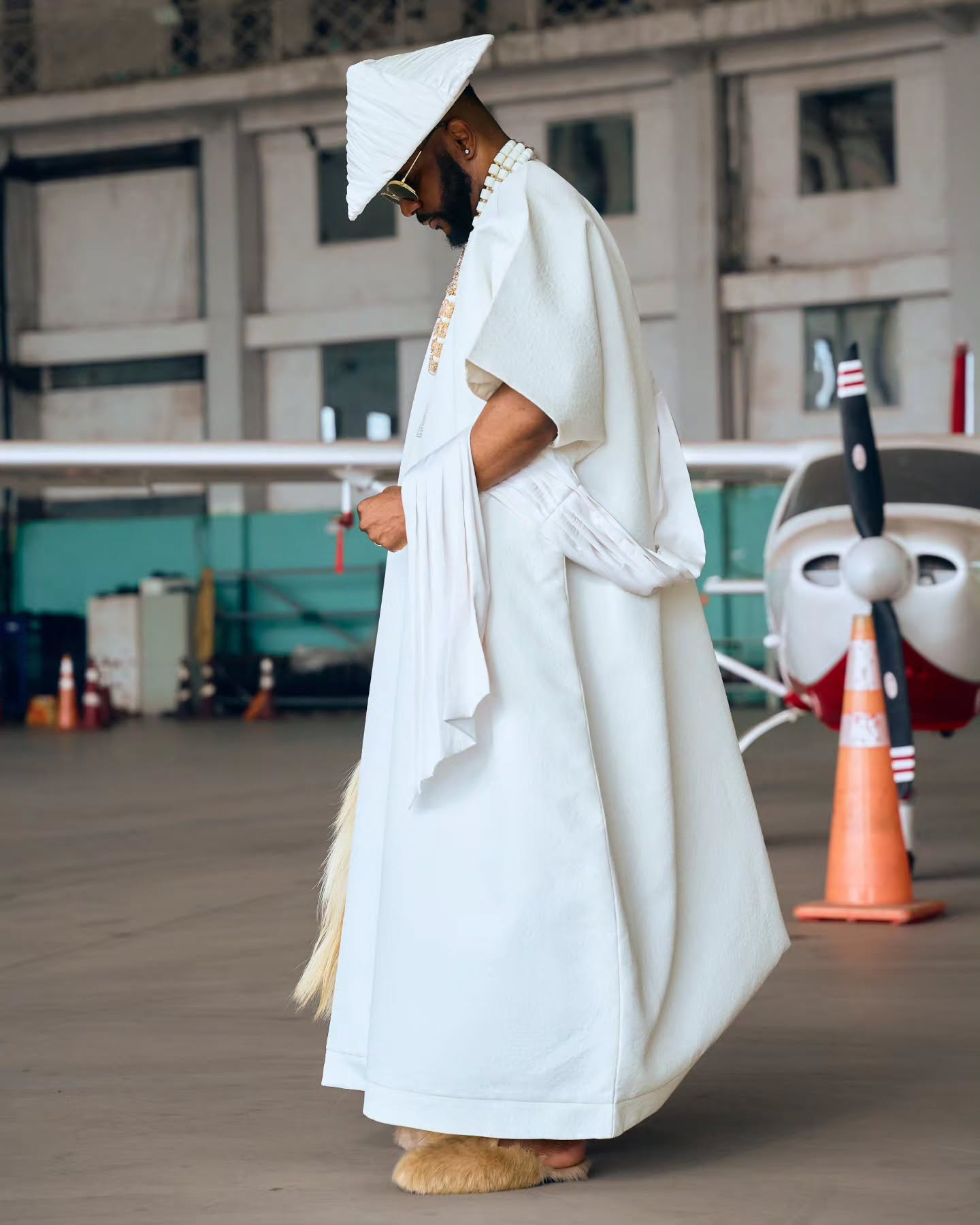 Ebuka Obi-Uchendu in a white Japanese-inspired robe and pleated Japanese hat standing in an aviation hangar next to a light aircraft.