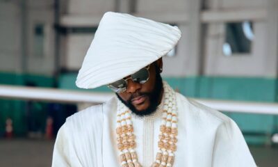 Ebuka Obi-Uchendu wearing a white pleated Japanese hat, gold-rimmed sunglasses, coral beads, and holding a blonde horsetail whip.