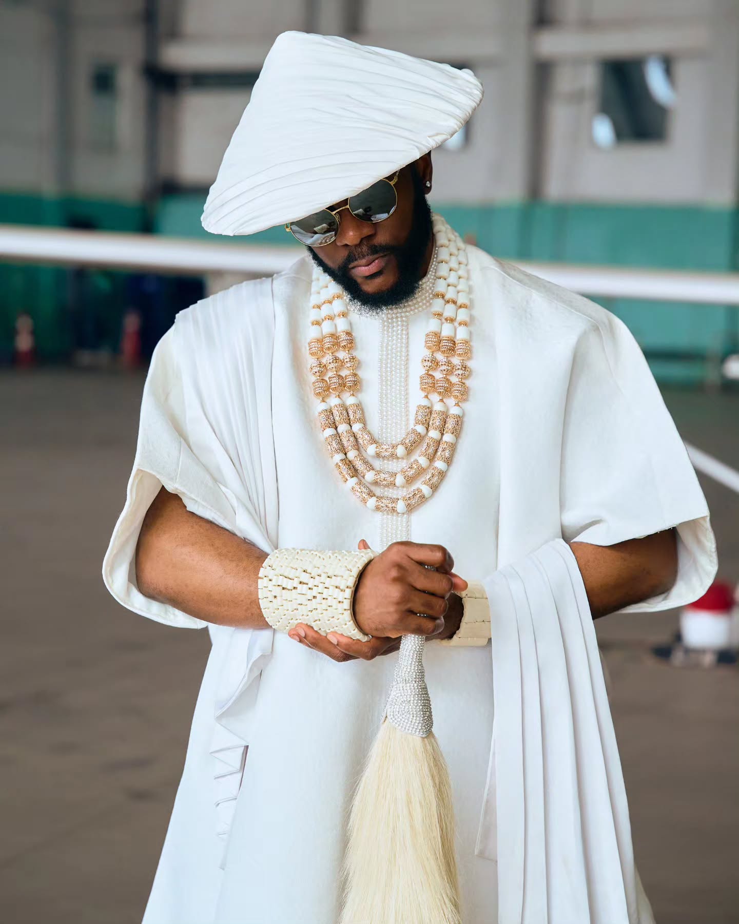 Ebuka Obi-Uchendu wearing a white pleated Japanese hat, gold-rimmed sunglasses, coral beads, and holding a blonde horsetail whip.