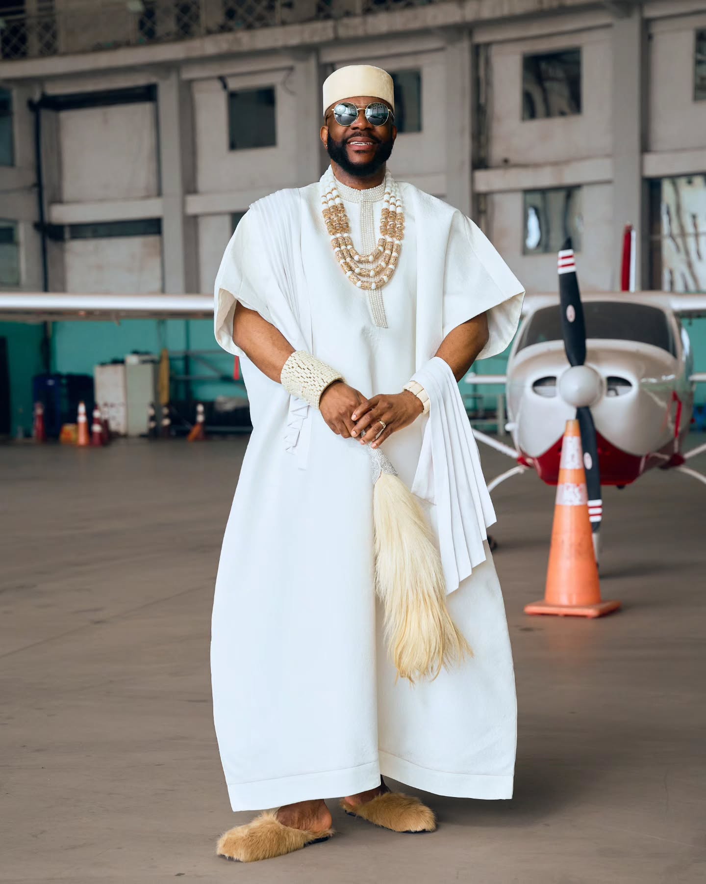 Ebuka Obi-Uchendu standing in a white Japanese-inspired Ugo Monye tunic set with sunglasses, cream cap, horsetail whip, and fur slippers.