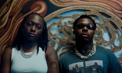 British-Nigerian artist Darkoo wearing a white top and gold chains standing beside a featured artist against a textured carved wall backdrop.