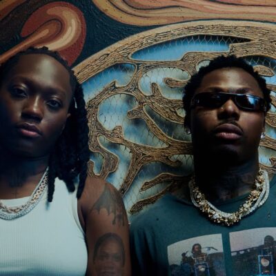 British-Nigerian artist Darkoo wearing a white top and gold chains standing beside a featured artist against a textured carved wall backdrop.
