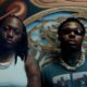 British-Nigerian artist Darkoo wearing a white top and gold chains standing beside a featured artist against a textured carved wall backdrop.
