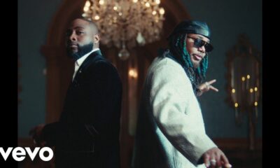 Davido in a black suit and Leon Thomas in a cream jacket standing back-to-back in the Tell Everybody music video.