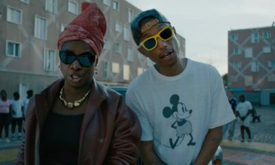 Angélique Kidjo in a burgundy leather jacket and pink headwrap standing beside Pharrell Williams wearing a Mickey Mouse t-shirt and yellow sunglasses in a street scene