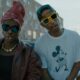 Angélique Kidjo in a burgundy leather jacket and pink headwrap standing beside Pharrell Williams wearing a Mickey Mouse t-shirt and yellow sunglasses in a street scene