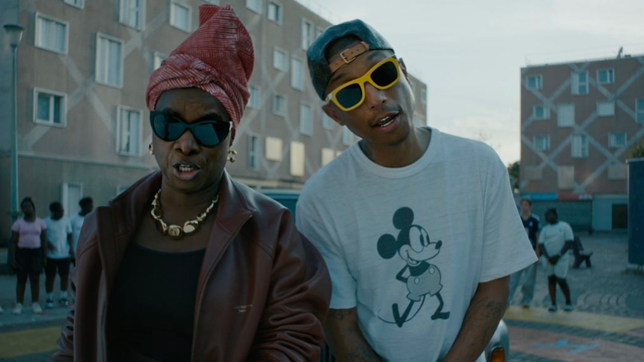 Angélique Kidjo in a burgundy leather jacket and pink headwrap standing beside Pharrell Williams wearing a Mickey Mouse t-shirt and yellow sunglasses in a street scene