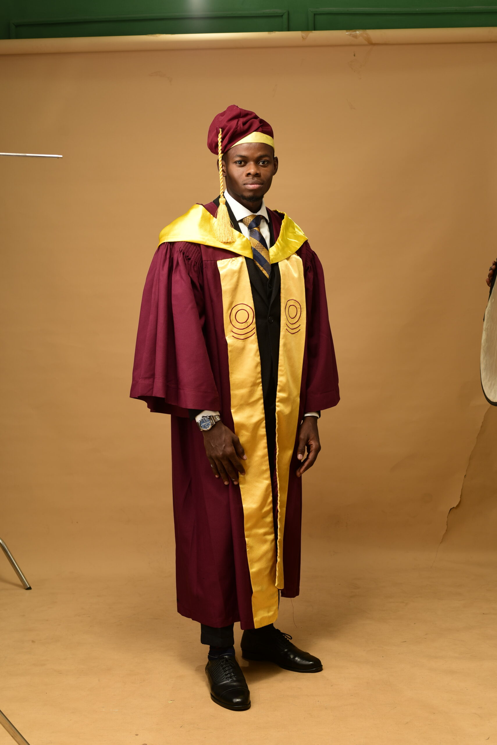 Haroun Adebakin in full University of Lagos graduation gown and cap standing on set.