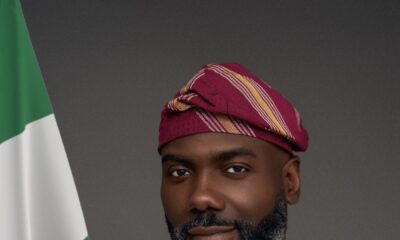 Olugbile Holloway, Director-General of the National Commission for Museums and Monuments (NCMM), wearing traditional white Agbada and a magenta striped Fila cap beside the Nigerian flag
