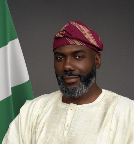 Olugbile Holloway, Director-General of the National Commission for Museums and Monuments (NCMM), wearing traditional white Agbada and a magenta striped Fila cap beside the Nigerian flag
