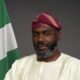 Olugbile Holloway, Director-General of the National Commission for Museums and Monuments (NCMM), wearing traditional white Agbada and a magenta striped Fila cap beside the Nigerian flag